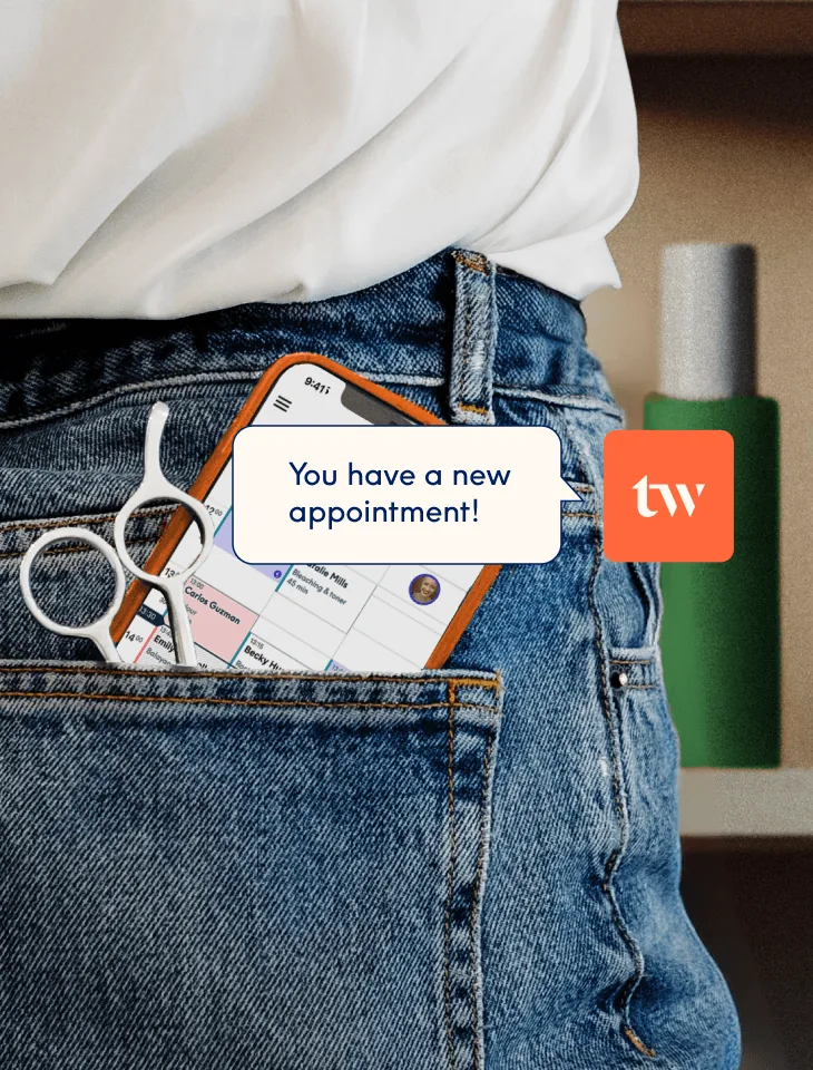 Un smartphone et une paire de ciseaux de coiffure glissés dans la poche arrière d'un jean, l'écran affichant l'agenda de réservations de l'application Treatwell avec une notification « vous avez un nouveau rendez-vous ! ».